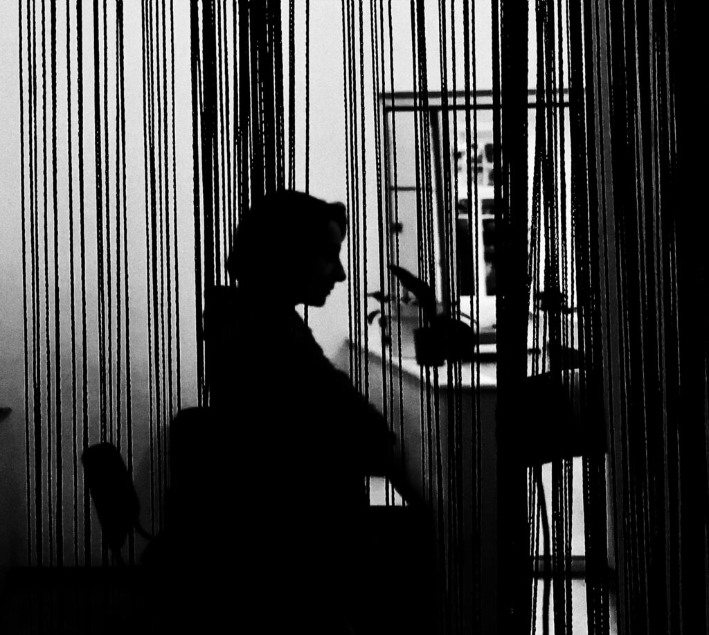 Black and white silhouette of a woman sitting behind a curtain indoors.